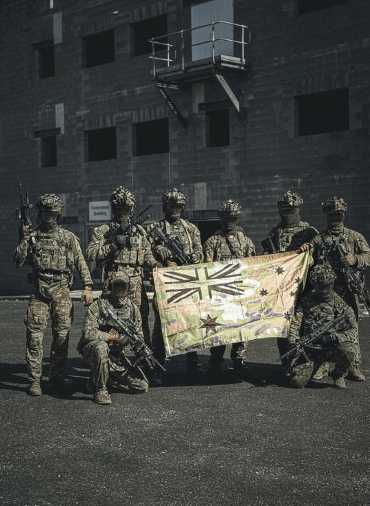 Group of soldiers in camouflage uniforms holding a flag in front of a building.